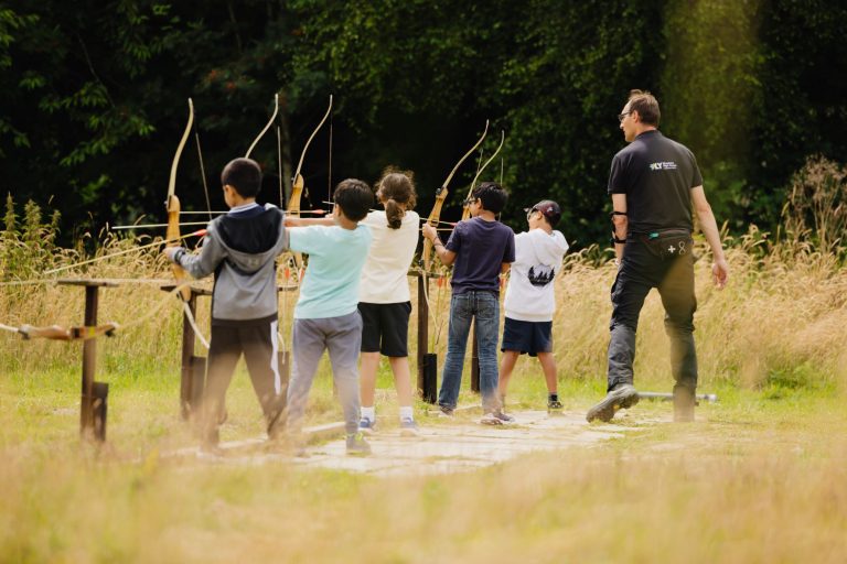 Outdoor instructor coaches children on archery range at Woodrow High House