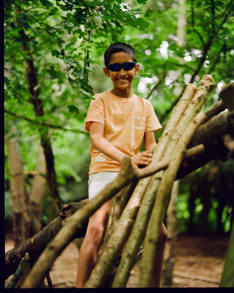 Boy with sunglasses smiling, sitting on top of shelter he's made in the woods